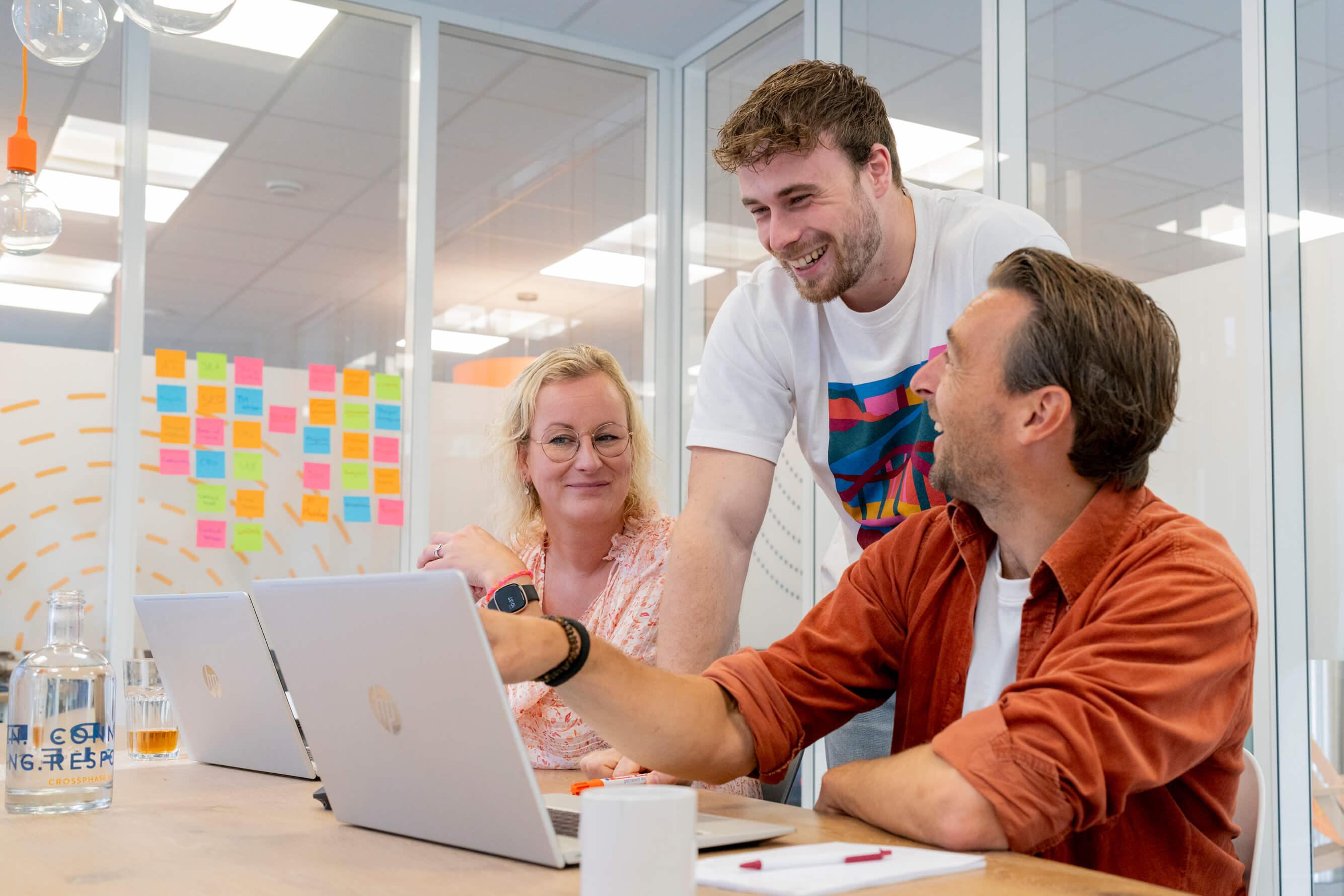 Drie collega's lachen en overleggen aan tafel met laptops