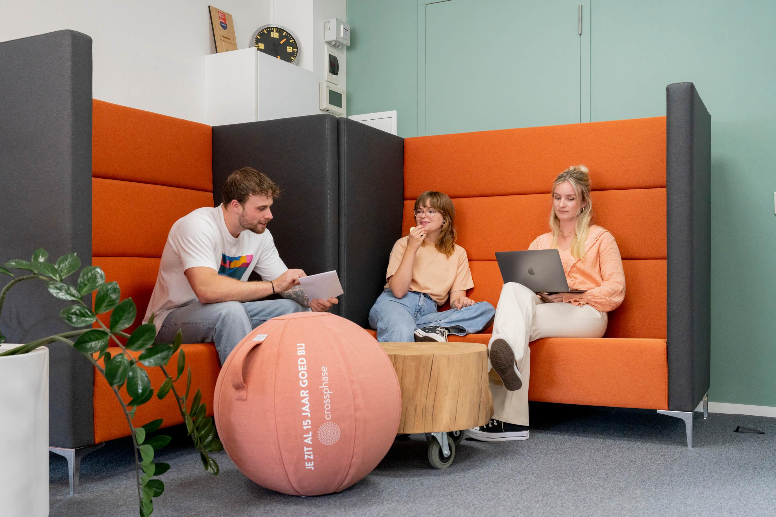 Drie collega's in gesprek in een oranje zithoek met laptop en notitieblok