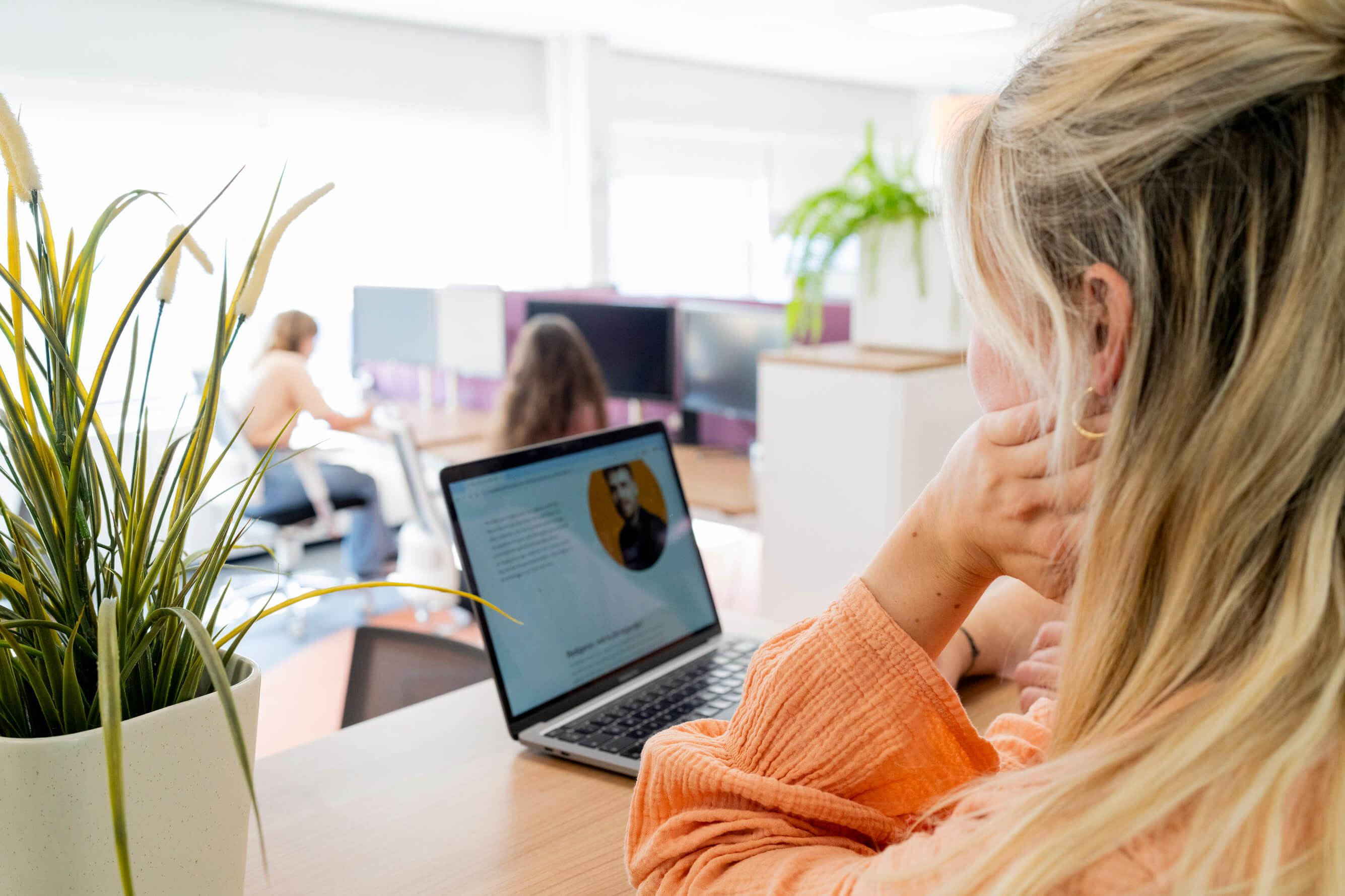Vrouw kijkt naar scherm van een laptop en op de achtergrond werken twee collega's